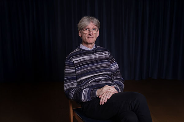 Älterer Mann mit grauen Haaren und Brille sitzt auf Stuhl vor dunklem Vorhang