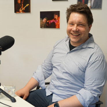 Mann mit hellblauem Hemd sitzt lächelnd vor Mikrofon an Tisch
