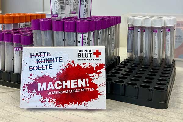 Gestell mit Blutprobenröhrchen und Schild Spende Blut beim Roten Kreuz Machen Gemeinsam Leben Retten
