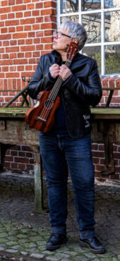 Person mit kurzer grauer Frisur und Brille hält eine Ukulele vor einer Backsteinwand.