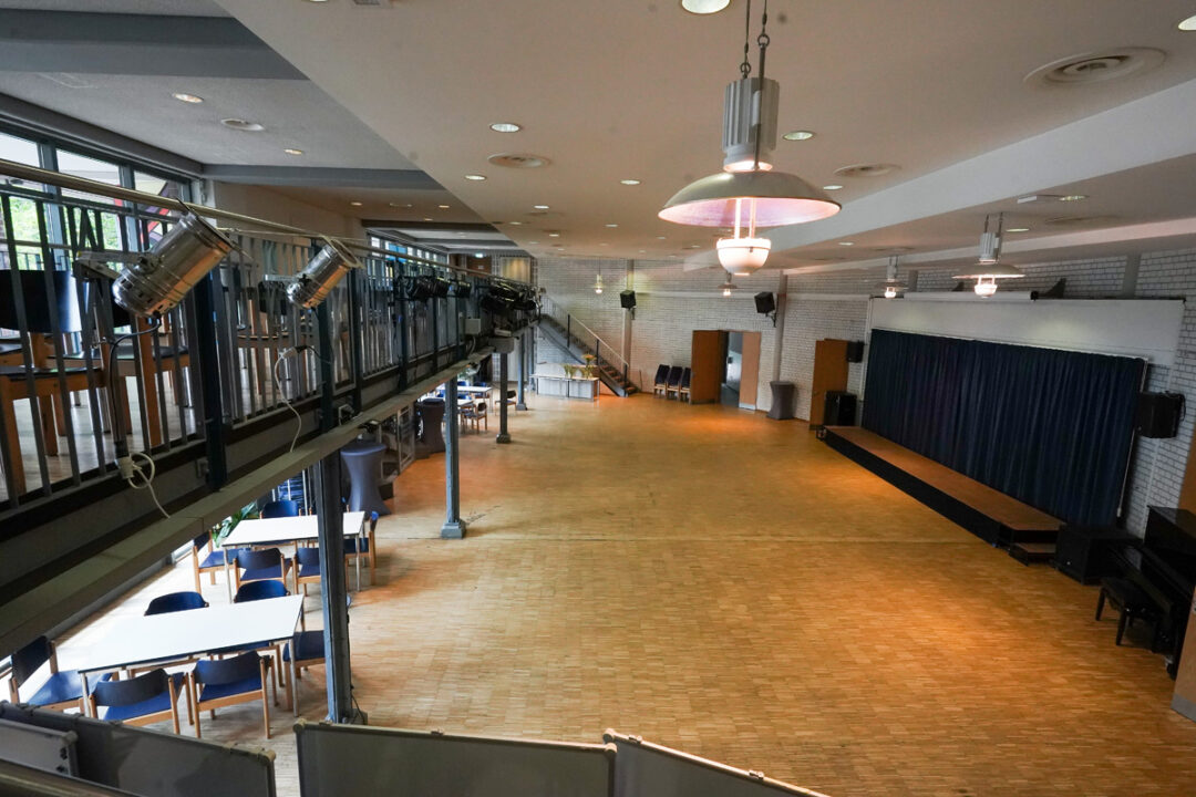 Großer Saal mit Parkettboden, Bühne mit Vorhang rechts und Galerie mit Beleuchtung links