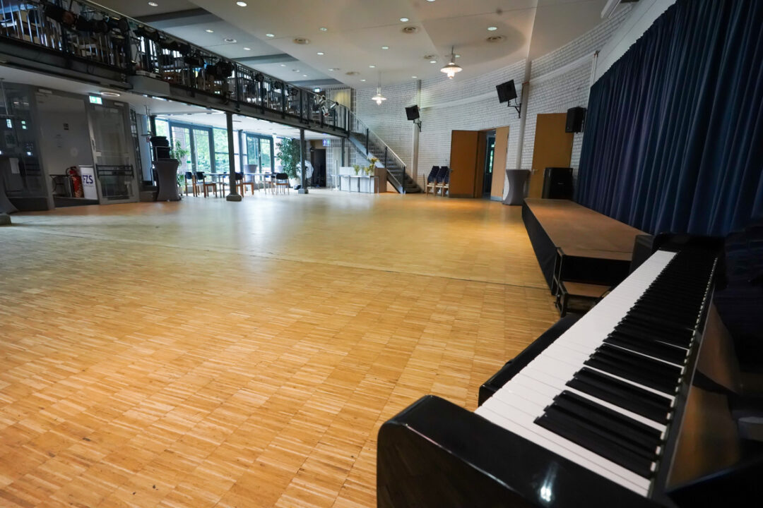 Großer Veranstaltungsraum mit Parkettboden, Bühne mit Vorhang und Klavier im Vordergrund
