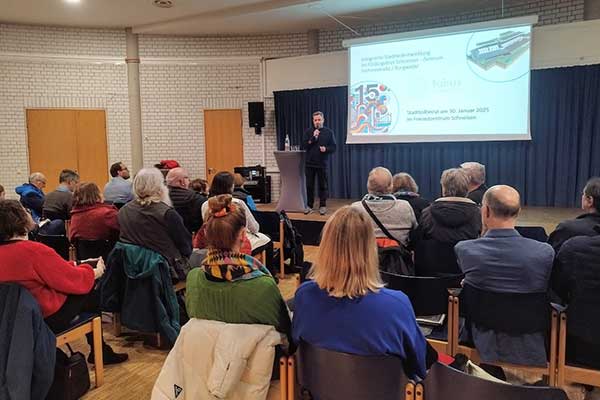 Vortragender steht auf Bühne vor Publikum, Präsentationsfolie im Hintergrund, Sitzreihen im Saal