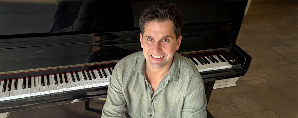 Erwachsener Mann sitzt vor Klavier, lächelt, Tastatur eines Flügels sichtbar im Hintergrund
