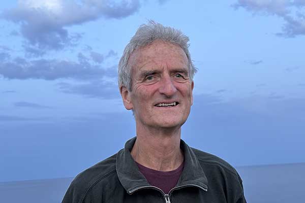 Älterer Mann mit grauen Haaren in dunkler Jacke vor bewölktem Himmel und Meer