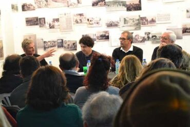 Podiumsdiskussion mit vier Personen vor Wand mit historischen Fotos, Publikum im Vordergrund