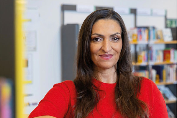 Person mit langem dunklem Haar und rotem Oberteil vor unscharfen Bücherregalen in einer Bibliothek oder Bücherei