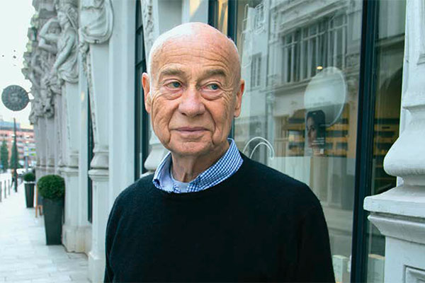 Älterer Mann mit Glatze und schwarzem Pullover steht vor einer historischen Gebäudefassade mit großen Fenstern.